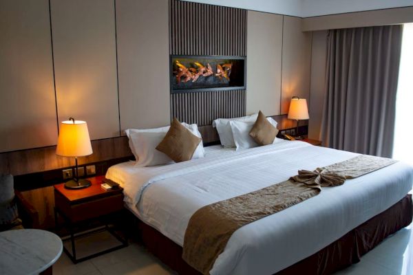 A modern hotel room with a large bed, two bedside lamps, cushions, a small table, and curtains. A decorative wall feature is above the bed.