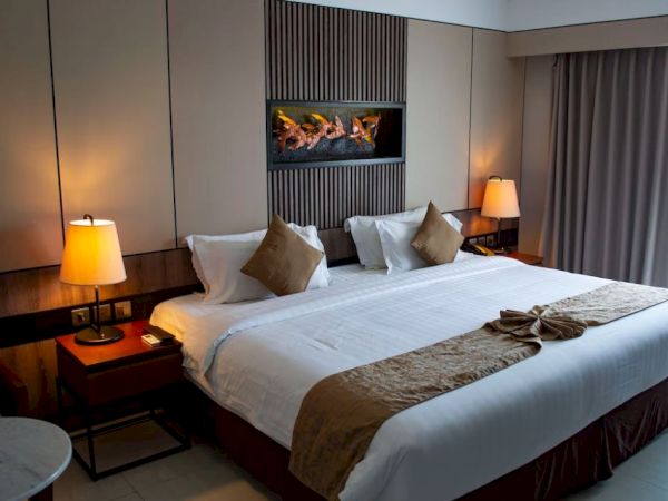 A modern hotel room with a large bed, two bedside lamps, cushions, a small table, and curtains. A decorative wall feature is above the bed.
