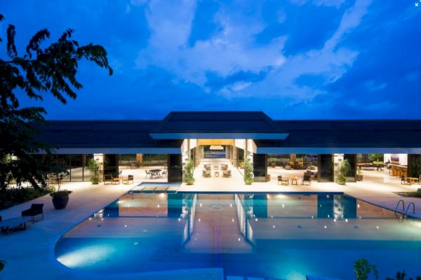 A modern building with an illuminated swimming pool at night, surrounded by seating and greenery under a blue sky.