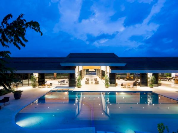 A modern building with an illuminated swimming pool at night, surrounded by seating and greenery under a blue sky.