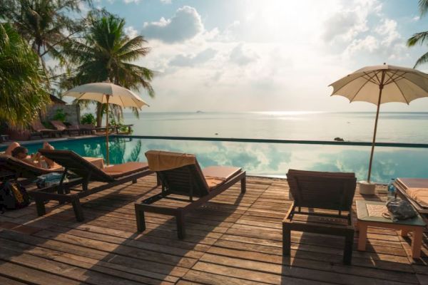 A serene tropical scene with lounge chairs, umbrellas, and a pool overlooking the ocean, surrounded by palm trees under a cloudy sky.