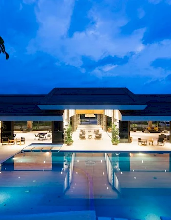 A luxurious outdoor swimming pool area at night, with a modern building and seating areas surrounded by bright lighting.