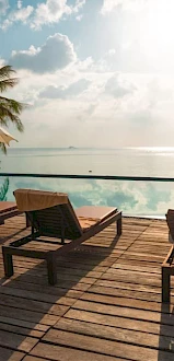 A serene poolside view with lounge chairs, umbrellas, and palm trees overlooking a calm body of water under a partly cloudy sky.