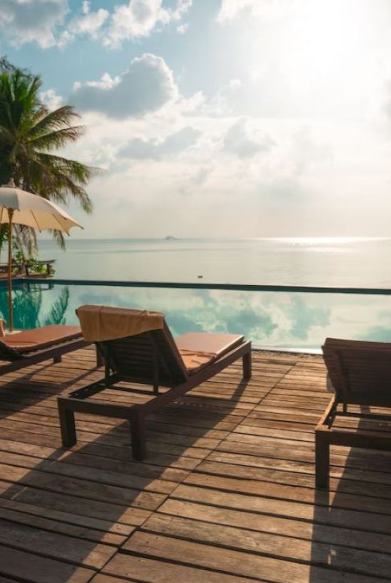 Wooden deck with lounge chairs and umbrellas by an infinity pool, overlooking a serene ocean view under a sunny sky.