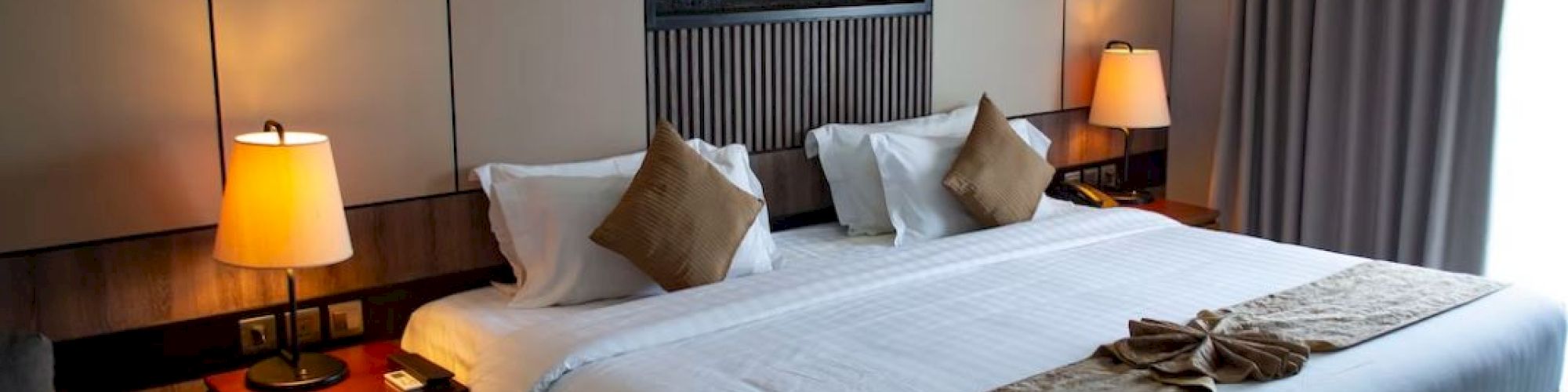 A cozy hotel room with a large bed, decorative pillows, bedside lamps, and wall art. Curtains cover a window to the right.
