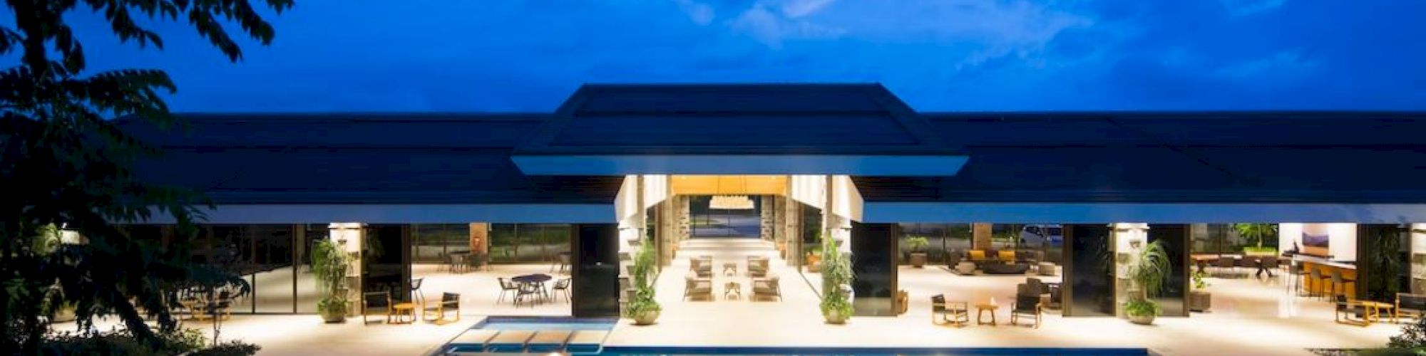 A luxurious resort with an illuminated pool and open-air pavilion under a twilight sky, surrounded by lush greenery and elegant architecture.