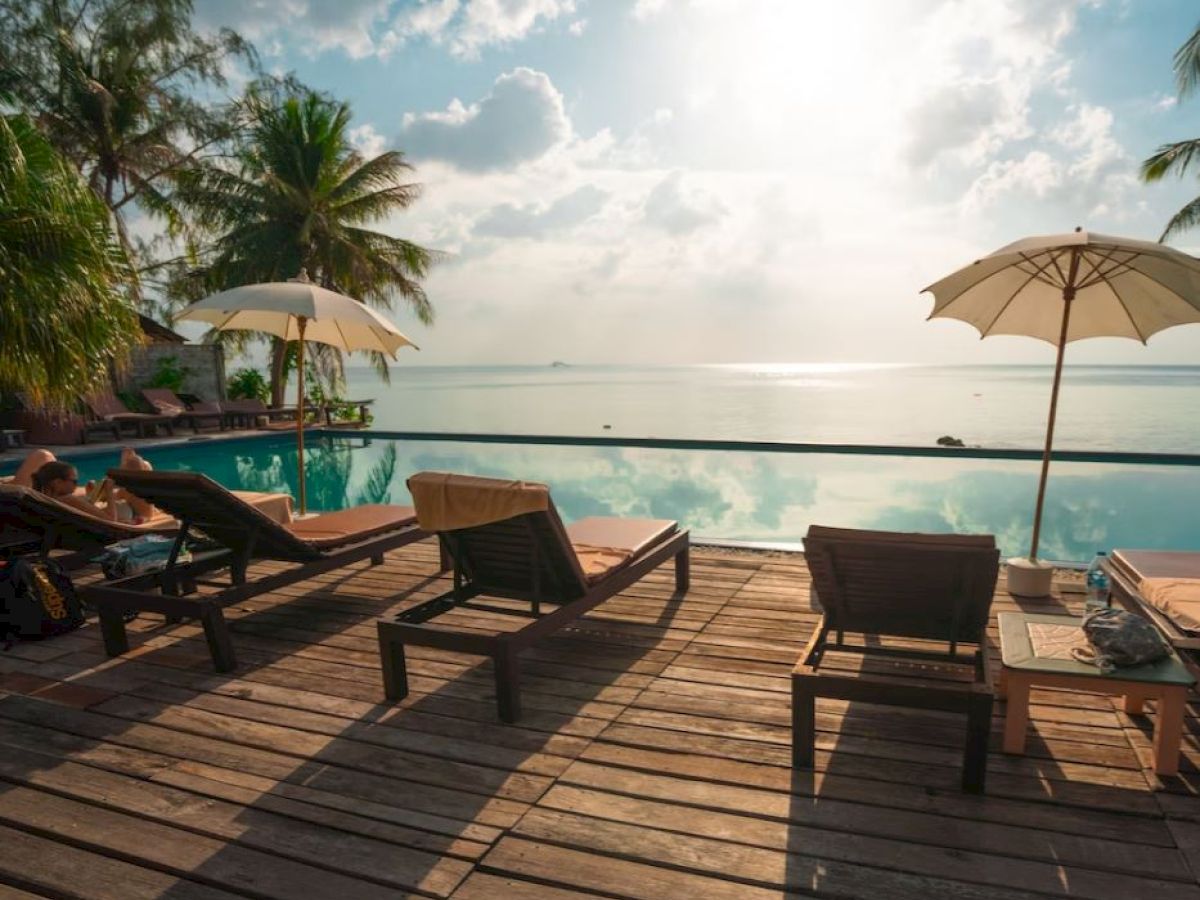 A serene poolside scene with lounge chairs, umbrellas, palm trees, and an ocean view under a bright, partly cloudy sky.