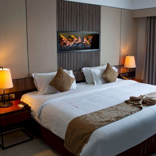 A well-decorated hotel room features a large bed with pillows, side tables with lamps, and a stylish wall with an artificial fireplace.