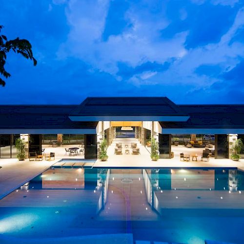 A luxurious building at dusk with a large, illuminated swimming pool reflecting the structure, surrounded by lounge chairs and greenery.