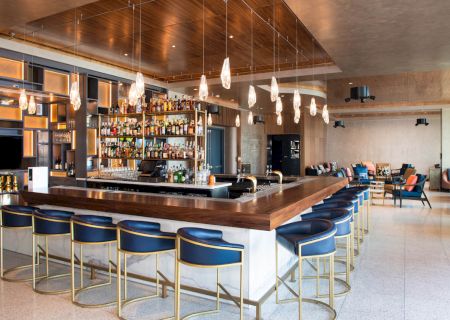 A modern bar with blue stools, hanging lights, and a stocked bar counter. The room has seating and a warm, stylish ambience.