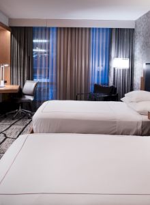 The image shows a modern hotel room with two beds, a TV, a desk, lamps, and a patterned floor and wall.
