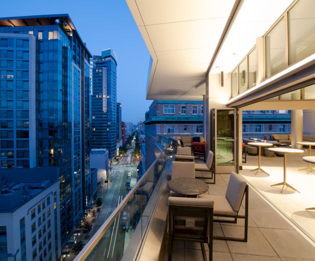 A modern cityscape at dusk with tall buildings, seen from a stylish open-air balcony with tables and chairs, partially indoors.