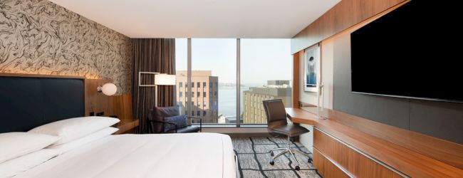 A modern hotel room features a large bed, wall-mounted TV, desk with chair, and a window with a cityscape view in the background.