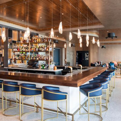 A chic bar with blue stools, pendant lights, and a well-stocked counter in a modern, spacious setting with seating for patrons.