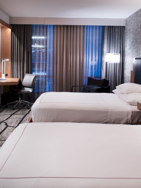 A modern hotel room with two beds, a TV, a desk, and stylish decor, featuring patterned wallpaper and a large window with curtains.
