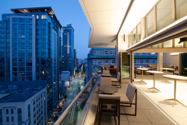 A modern cityscape at dusk with a rooftop terrace featuring tables and chairs, overlooking a street lined with tall buildings.