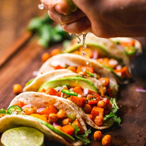 A hand squeezes lime juice over tacos filled with chickpeas, diced veggies, and avocado slices on a wooden board.