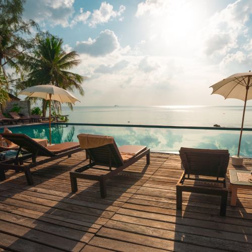 A serene beach scene with lounge chairs, umbrellas, and a pool overlooking the ocean under a bright sky and palm trees in the background.