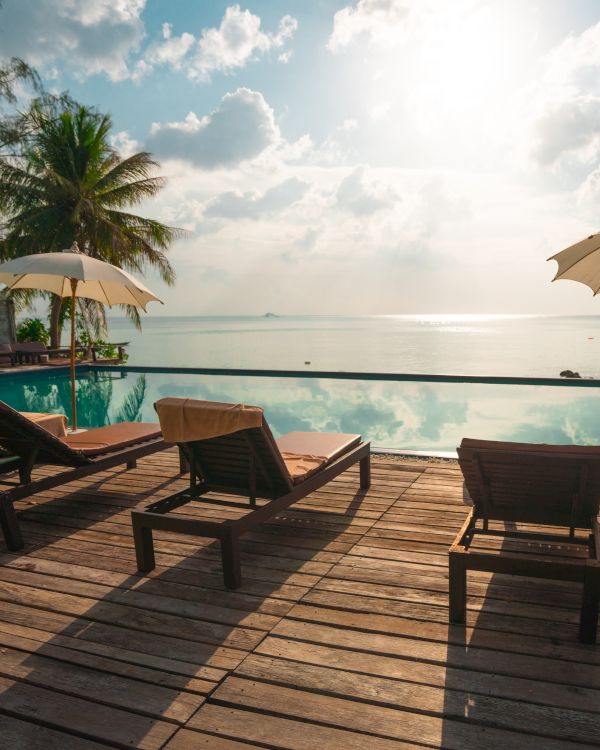 A serene beach scene with lounge chairs, umbrellas, and a pool overlooking the ocean under a bright sky and palm trees in the background.