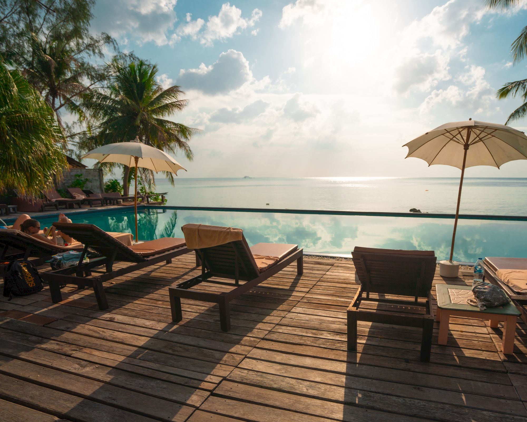 A beachside pool with lounge chairs and umbrellas, overlooking the ocean under a bright sky with clouds and palm trees.