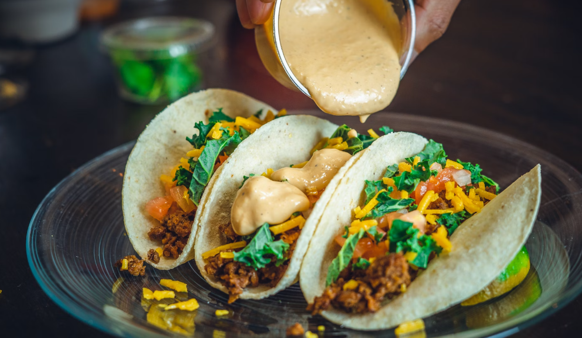 Three tacos filled with greens, cheese, and a creamy sauce being poured, are arranged on a plate with a lime wedge.