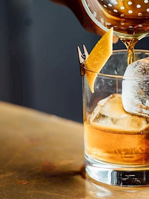 A cocktail is being poured into a glass with ice and an orange twist garnish on a metal surface.