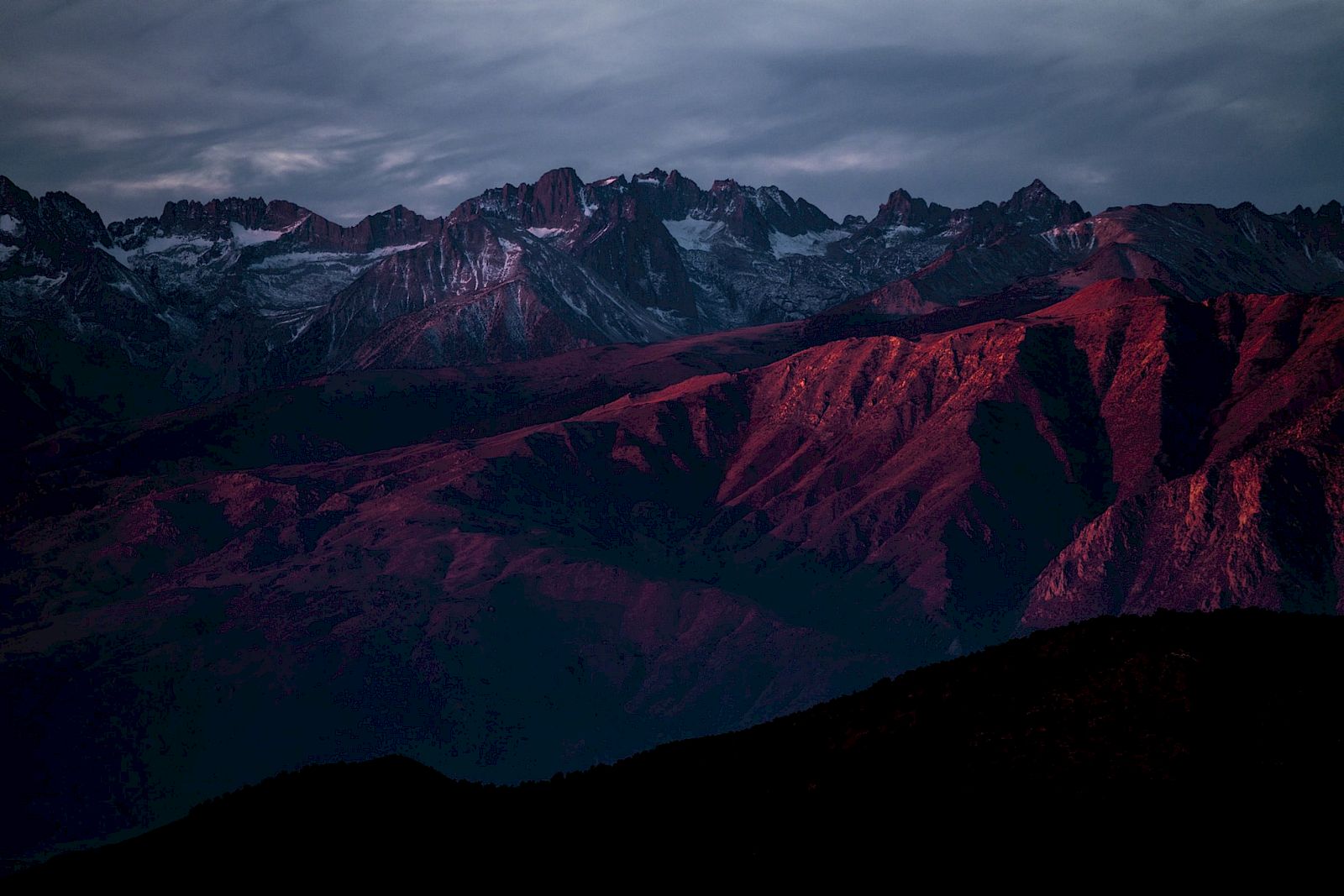 A mountain range is illuminated by a reddish sunset, casting dramatic shadows and highlights across the rocky landscape.
