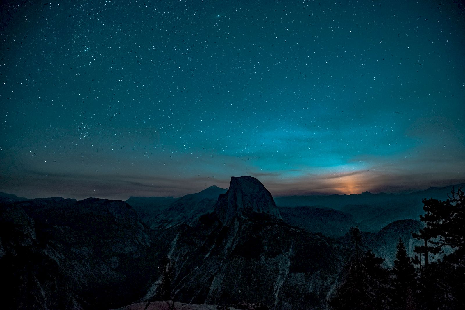 A starry night sky over a mountain landscape with a glowing horizon. Trees are silhouetted in the foreground, creating a serene scene.