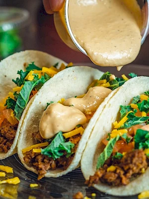 Three tacos filled with ground meat, cheese, lettuce, and tomato, topped with sauce being poured from a small container on a plate.