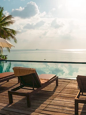 Poolside view with lounge chairs, umbrellas, and palm trees overlooking a calm sea at sunset. Peaceful tropical setting.