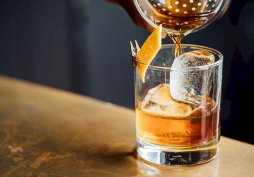 A hand is pouring a brown liquid into a glass with a large ice cube and a twist of lemon peel, on a wooden surface.