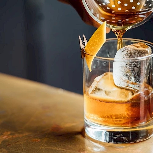 A hand is pouring a brown liquid into a glass with a large ice cube and a twist of lemon peel, on a wooden surface.