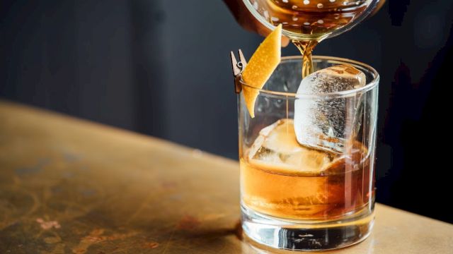 A hand is pouring a brown liquid into a glass with a large ice cube and a twist of lemon peel, on a wooden surface.