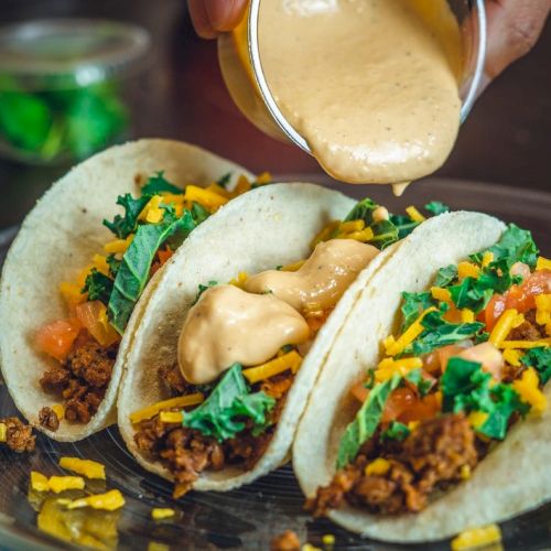 Three tacos filled with ground meat, cheese, tomatoes, and greens, topped with sauce being poured from a cup.