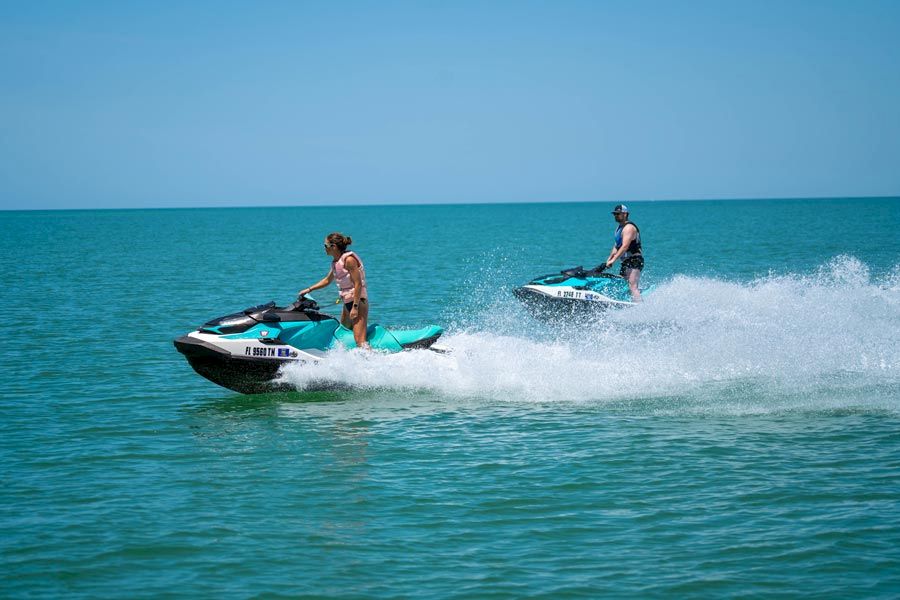 Two people are riding jet skis on a clear blue sea, creating splashes as they speed across the water under a sunny sky.