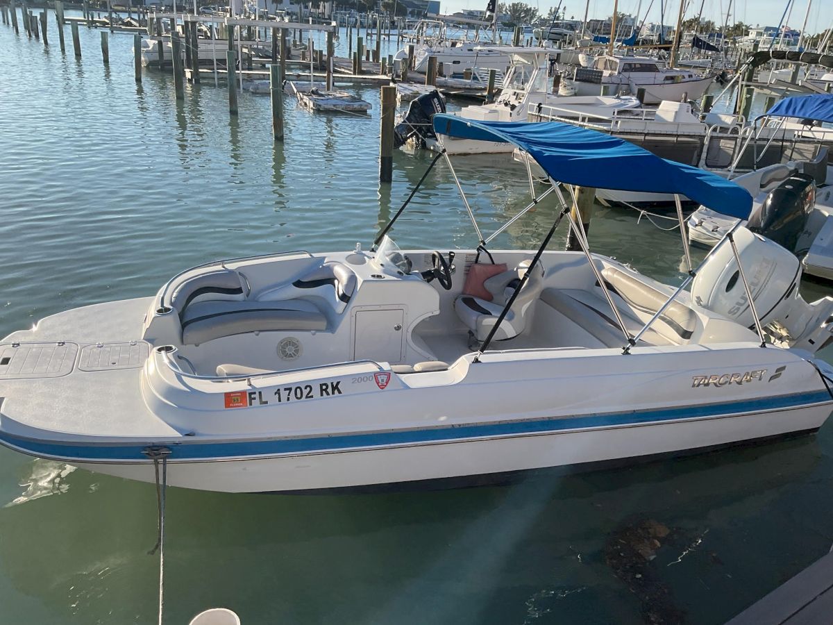 A small white motorboat docked at a marina, with a blue canopy, calm water, and other boats in the background.