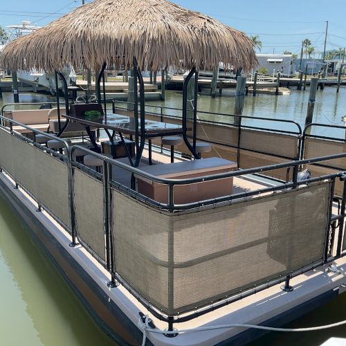 A floating tiki bar with a thatched roof and seating is docked in a marina. The structure is enclosed with railings.
