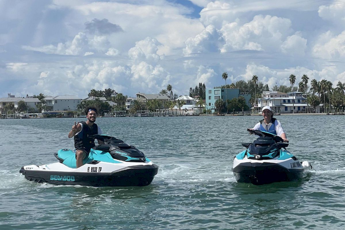Two people are riding jet skis on a body of water with a backdrop of buildings and palm trees under a partly cloudy sky.