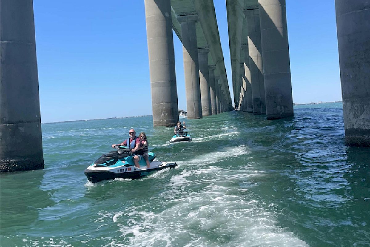 People riding jet skis under a large concrete bridge over a body of water, creating waves as they move away.