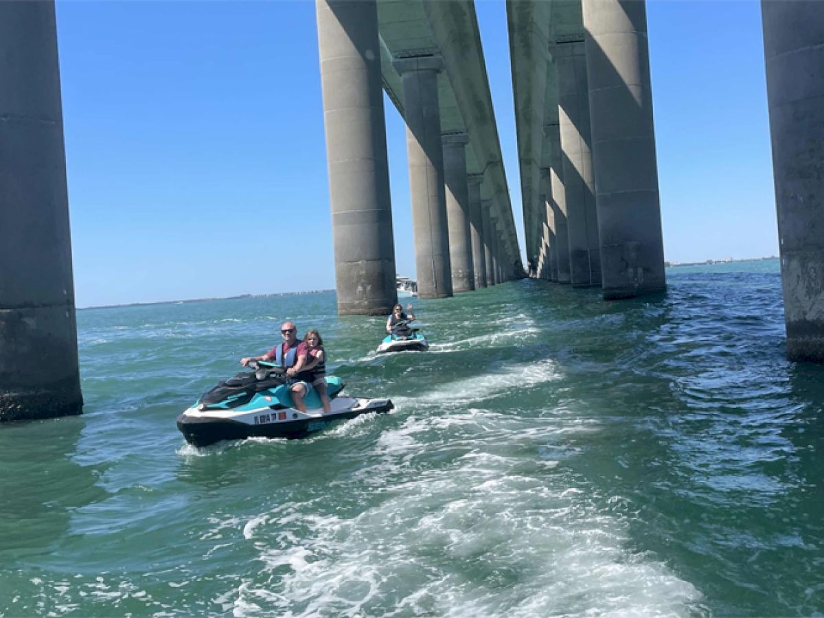 People riding jet skis under a large concrete bridge over a body of water, creating waves as they move away.
