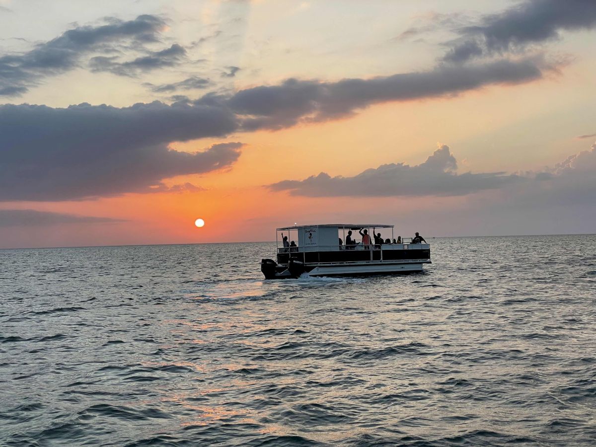 A boat cruises on calm ocean at sunset, passengers waving as the sun sinks behind clouds.