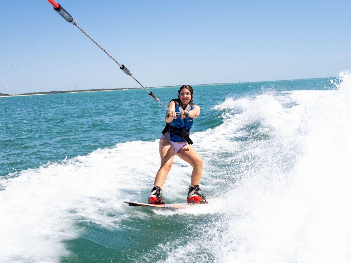 A woman kiteboards across choppy blue water, harness on, holding the control bar as waves splash behind her.