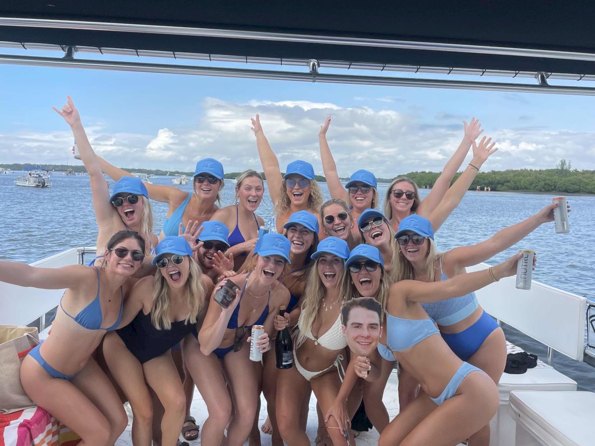 A cheering group of friends in blue hats and swimsuits on a boat, arms up, celebrating on a sunny lake with drinks in hand.