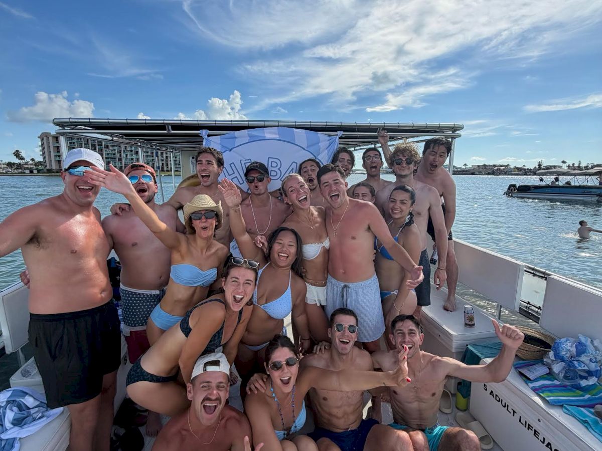 A cheering group of friends in swimsuits pose on a boat on a sunny ocean day with a &ldquo;...BAB...&rdquo; banner behind them.
