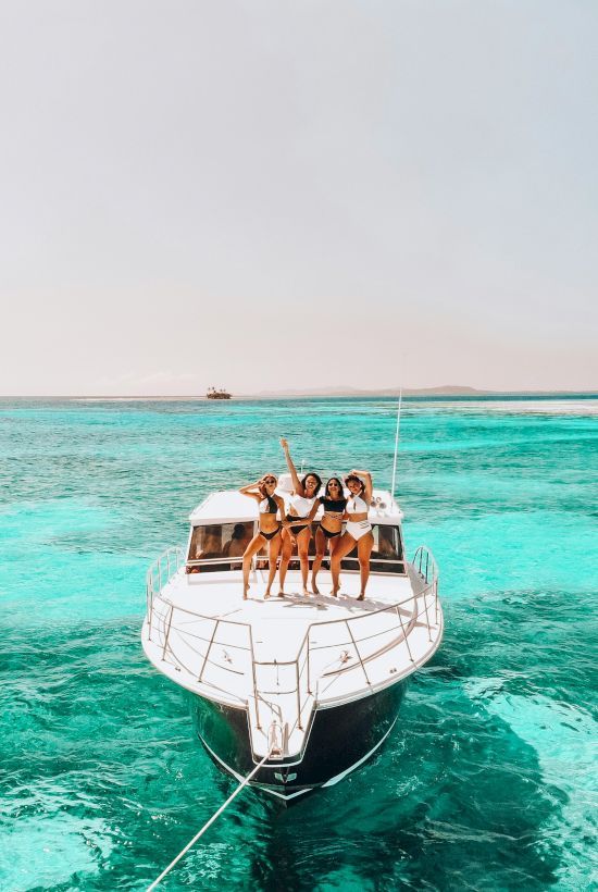 A group of people in swimwear are posing and celebrating on the bow of a boat in clear turquoise waters.