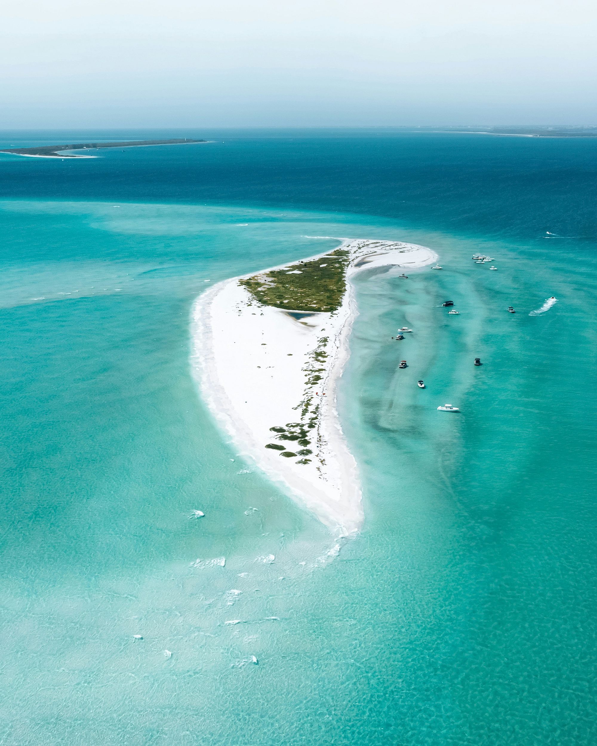 An aerial view of a small, narrow island surrounded by clear turquoise water and a few boats nearby.