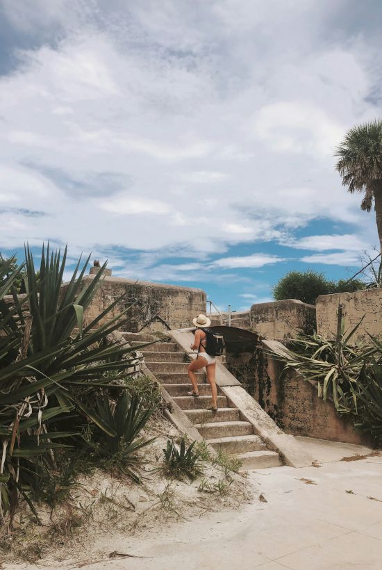 A person in a swimsuit and hat ascends stone steps surrounded by greenery, with a blue sky and palm tree in the background.