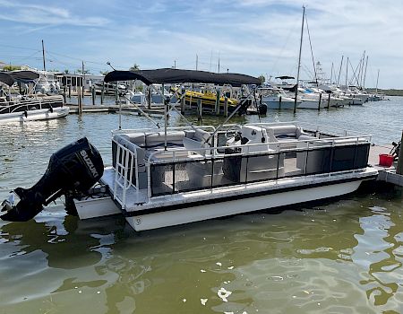 A white pontoon boat with a black canopy is docked at a marina, surrounded by other boats under cloudy skies.