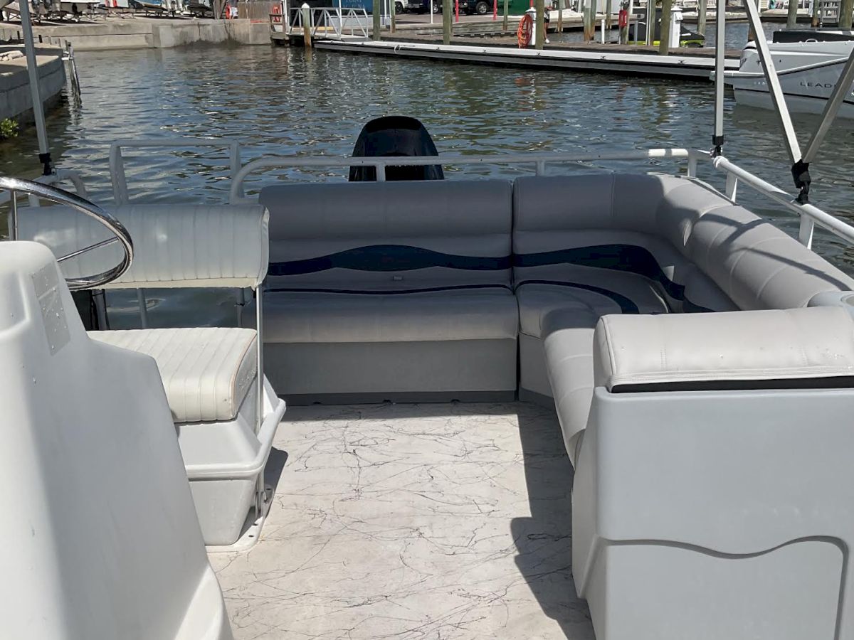 A pontoon boat cockpit with white seating looks over a marina canal toward docks, houses, and parked boats on a sunny day.