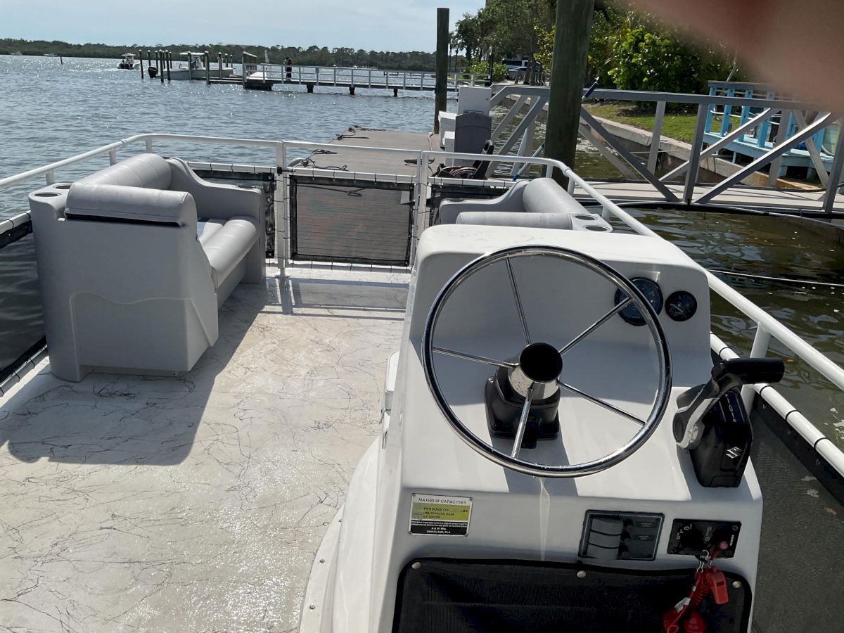 A boat cockpit with steering wheel and seats, docked by a calm lake with trees and piers in the background.